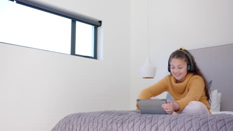 Happy-biracial-girl-sitting-on-bed,-using-tablet-and-headphones-in-sunny-bedroom,-copy-space