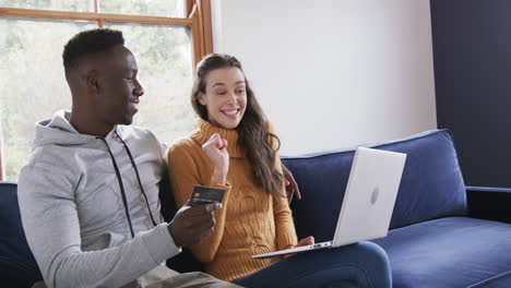 Happy-diverse-couple-sitting-on-sofa,-using-laptop-and-credit-card-in-home,copy-space