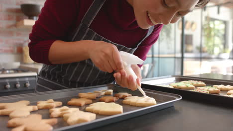 Happy-biracial-woman-in-apron-decorating-christmas-cookies-in-kitchen,-slow-motion