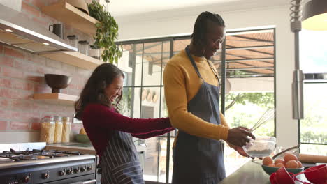 Happy-diverse-couple-in-aprons-baking-in-sunny-kitchen,-slow-motion