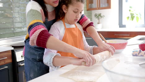 Happy-biracial-mother-and-daughter-rolling-out-dough-and-smiling-in-sunny-kitchen