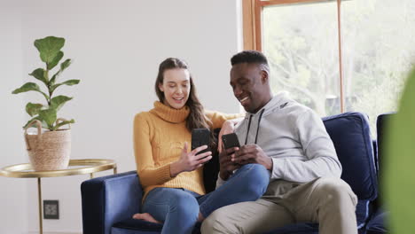 Happy-diverse-couple-sitting-on-sofa,-using-smartphone-and-credit-card-in-home,copy-space