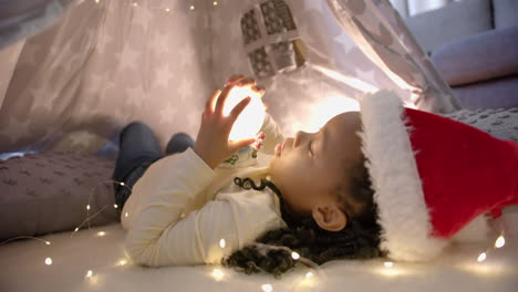 Happy-african-american-girl-in-christmas-hat-holding-glowing-snow-globe-in-blanket-tent,-slow-motion