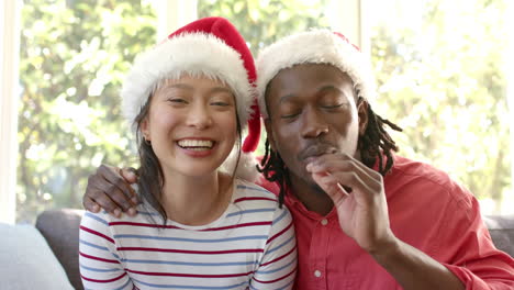 Glückliches,-Vielfältiges-Paar-In-Weihnachtsmützen-Mit-Videoanruf-Im-Sonnigen-Wohnzimmer,-Zeitlupe