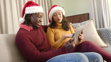 Glückliches,-Vielfältiges-Paar-In-Weihnachtsmützen-Mit-Tablet-Auf-Der-Couch-Zu-Hause,-Zeitlupe