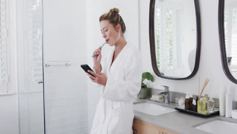 Biracial-woman-brushing-teeth-using-smartphone-in-bathroom,-slow-motion