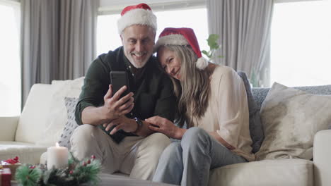 Middle-aged-caucasian-couple-in-santa-hats-on-smartphone-chat-at-christmas-at-home,-slow-motion