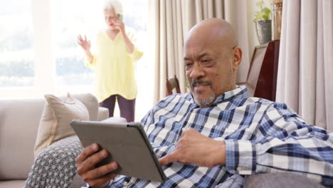 Glückliches,-Vielfältiges-Seniorenpaar-Mit-Smartphone-Und-Tablet-Im-Sonnigen-Wohnzimmer,-Zeitlupe