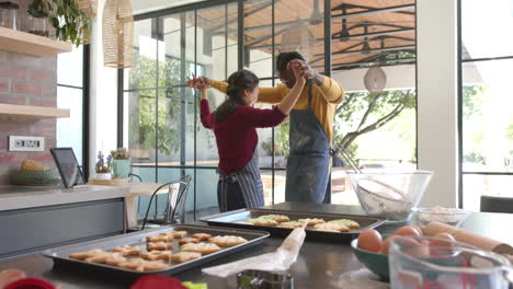 Happy-diverse-couple-in-aprons-baking-cookies-and-dancing-in-sunny-kitchen,-copy-space,-slow-motion