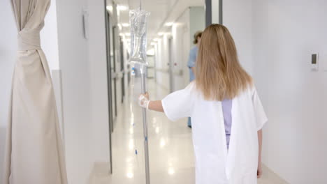 Rear-view-of-caucasian-girl-patient-walking-with-drip-stand-in-hospital,-copy-space,-slow-motion