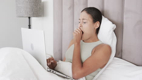 Biracial-woman-sitting-on-bed-using-laptop-and-yawning,-slow-motion