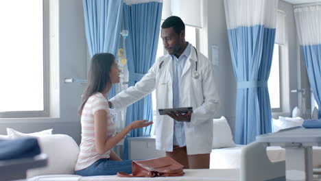 Diverse-male-doctor-using-tablet-and-female-patient,-talking-in-hospital-ward,-slow-motion