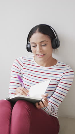 Vertical-video-of-portrait-of-happy-biracial-woman-using-headphones,-taking-notes,-slow-motion