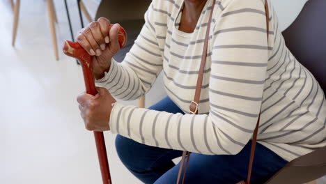 Midsection-of-african-american-senior-woman-with-cane-sitting-in-hospital-waiting-room,-slow-motion