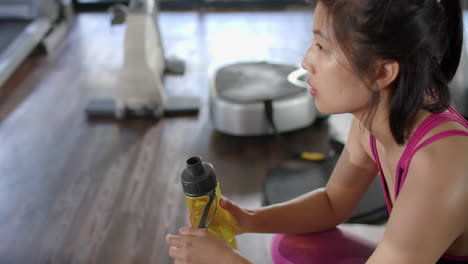 Tired-asian-woman-drinking-from-water-bottle-in-sunny-home-gym,-slow-motion