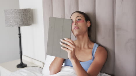 Biracial-woman-with-under-eye-patches-sitting-on-bed-using-tablet,-slow-motion