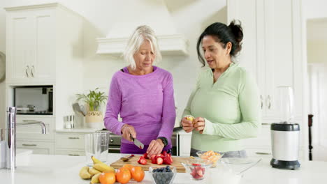 Two-happy-diverse-senior-women-chopping-fruits-and-laughing-in-kitchen,-slow-motion