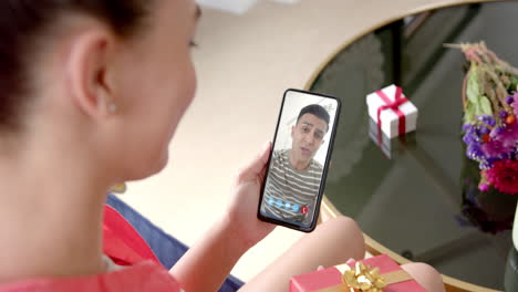 Biracial-woman-holding-smartphone-with-biracial-man-with-gift-on-screen-with-gift-on-desk