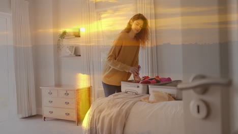 Animation-of-happy-caucasian-woman-packing-suitcase-in-bedroom-over-sunset-sky
