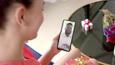Caucasian-woman-holding-gift-and-smartphone-with-african-emerican-man-on-screen
