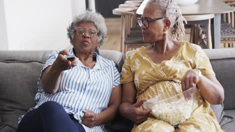 Glückliche-ältere-Afroamerikanische-Freundinnen-Auf-Der-Couch,-Die-Popcorn-Essen,-Mit-Der-TV-Fernbedienung,-Zeitlupe