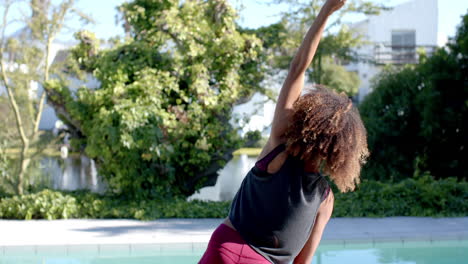 Happy-biracial-woman-practicing-yoga-stretching-by-pool-in-sunny-garden,-slow-motion,-copy-space