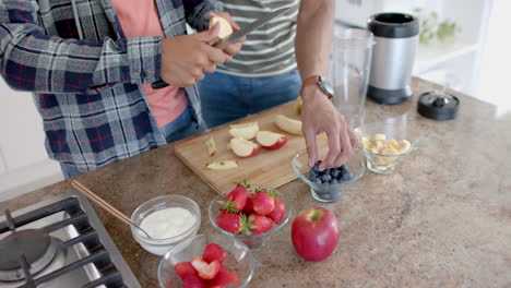Happy-diverse-gay-male-couple-preparing-fruit-for-smoothie-in-kitchen,-copy-space,-slow-motion