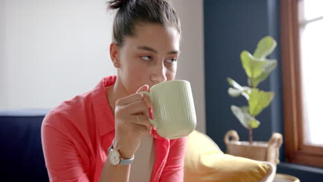 Thoughtful-biracial-teenage-girl-sitting-at-home-drinking-tea,-copy-space,-slow-motion