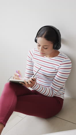 Vertical-video-of-biracial-woman-sitting-on-stairs-using-headphones-and-taking-notes,-slow-motion