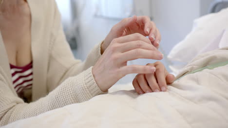 Midsection-of-caucasian-mother-holding-hand-of-daughter-patient-lying-in-hospital-bed,-slow-motion