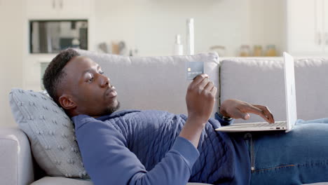 Glücklicher-Afroamerikanischer-Mann,-Der-Im-Sonnigen-Wohnzimmer-Liegt-Und-Laptop-Und-Kreditkarte-Benutzt,-Zeitlupe