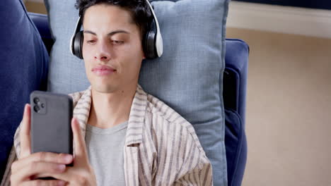 Glücklicher-Biracial-Mann-Mit-Kopfhörern,-Der-Auf-Der-Couch-Liegt-Und-Smartphone,-Kopierraum-Und-Zeitlupe-Verwendet