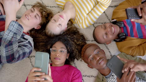 Happy-diverse-group-of-teenage-friends-using-smartphones-and-lying-on-carpet-at-home,-slow-motion