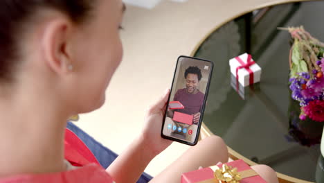 Biracial-woman-holding-smartphone-with-african-american-man-with-gift-on-screen-with-gift-on-desk