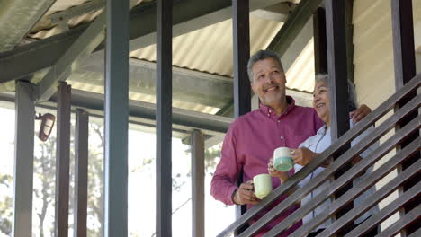 Happy-biracial-senior-couple-embracing-on-balcony,-talking-and-drinking-tea