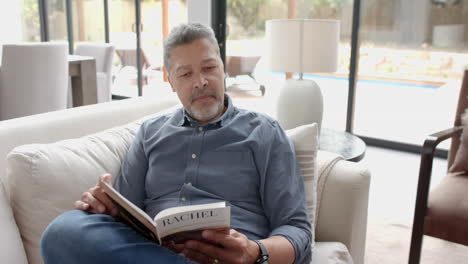 Focused-senior-biracial-man-sitting-on-couch-reading-book-in-living-room-at-home,-slow-motion