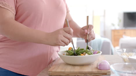 Happy-plus-size-biracial-woman-tossing-bowl-of-feta-vegetable-salad-in-kitchen,-slow-motion