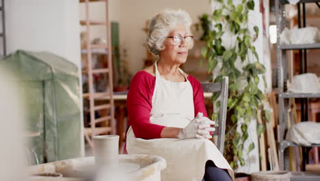 Happy-senior-biracial-female-potter-with-gray-hair-resting-smiling-in-pottery-studio,-slow-motion