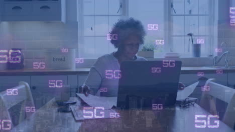 Animation-of-5g-text-repeated-over-senior-african-american-woman-using-laptop-in-kitchen