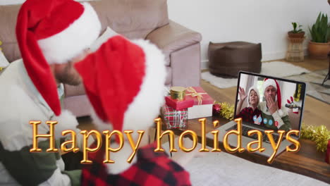 Animation-of-happy-holidays-text-over-friends-in-santa-hats-on-laptop-call-at-christmas