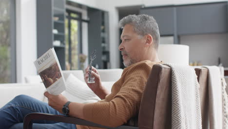Focused-senior-biracial-man-sitting-in-armchair-reading-book-in-living-room,-slow-motion