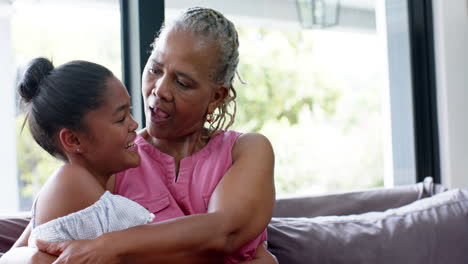 Happy-african-american-granddaughter-and-grandmother-embracing-on-sofa,-copy-space,-slow-motion