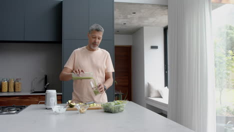 Happy-senior-biracial-man-pouring-healthy-smoothie-into-glass-in-kitchen-at-home,-slow-motion