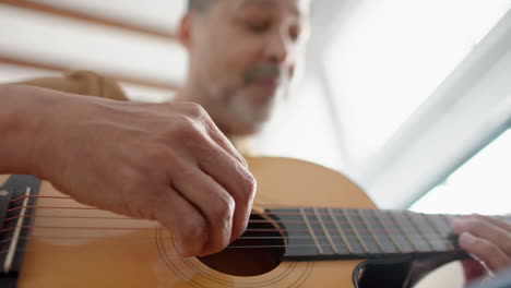 Low-angle-of-senior-biracial-man-sitting-playing-guitar-at-home,-selective-focus,-slow-motion