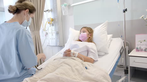 Caucasian-female-doctor-using-stethoscope-and-girl-patient-in-bed-in-face-masks,-slow-motion