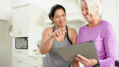 Two-happy-diverse-senior-women-using-tablet-and-laughing-in-kitchen,-slow-motion
