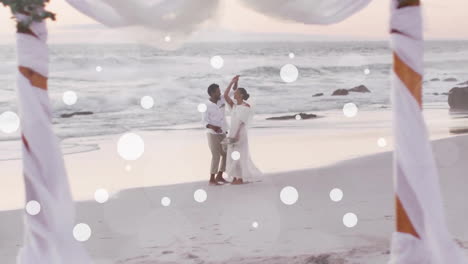 Animation-of-white-spots-over-african-american-bride-and-groom-dancing-on-beach-at-wedding