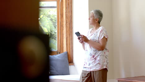 Nachdenkliche-ältere-Frau-Mit-Gemischter-Abstammung,-Die-Zu-Hause-Am-Fenster-Ihr-Smartphone-Benutzt,-Zeitlupe
