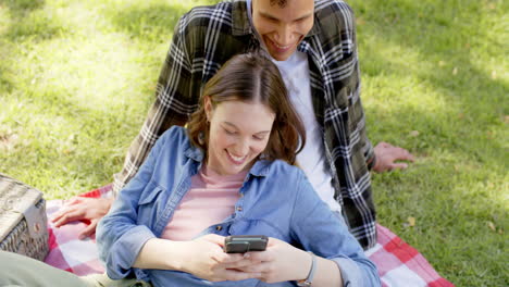 Happy-diverse-couple-having-picnic-and-using-smartphone-in-sunny-garden,-in-slow-motion