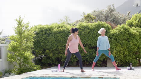 Two-happy-diverse-senior-women-practising-yoga-by-pool-in-sunny-garden,-slow-motion,-copy-space